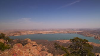 Aerial view from the summit of Mount Scott looking down over the blue-green waters of Lake Lawtonka, with reddish lichen-covered granite boulders and juniper in the foreground and Oklahoma's plains and distant wind turbines stretching to a hazy horizon.